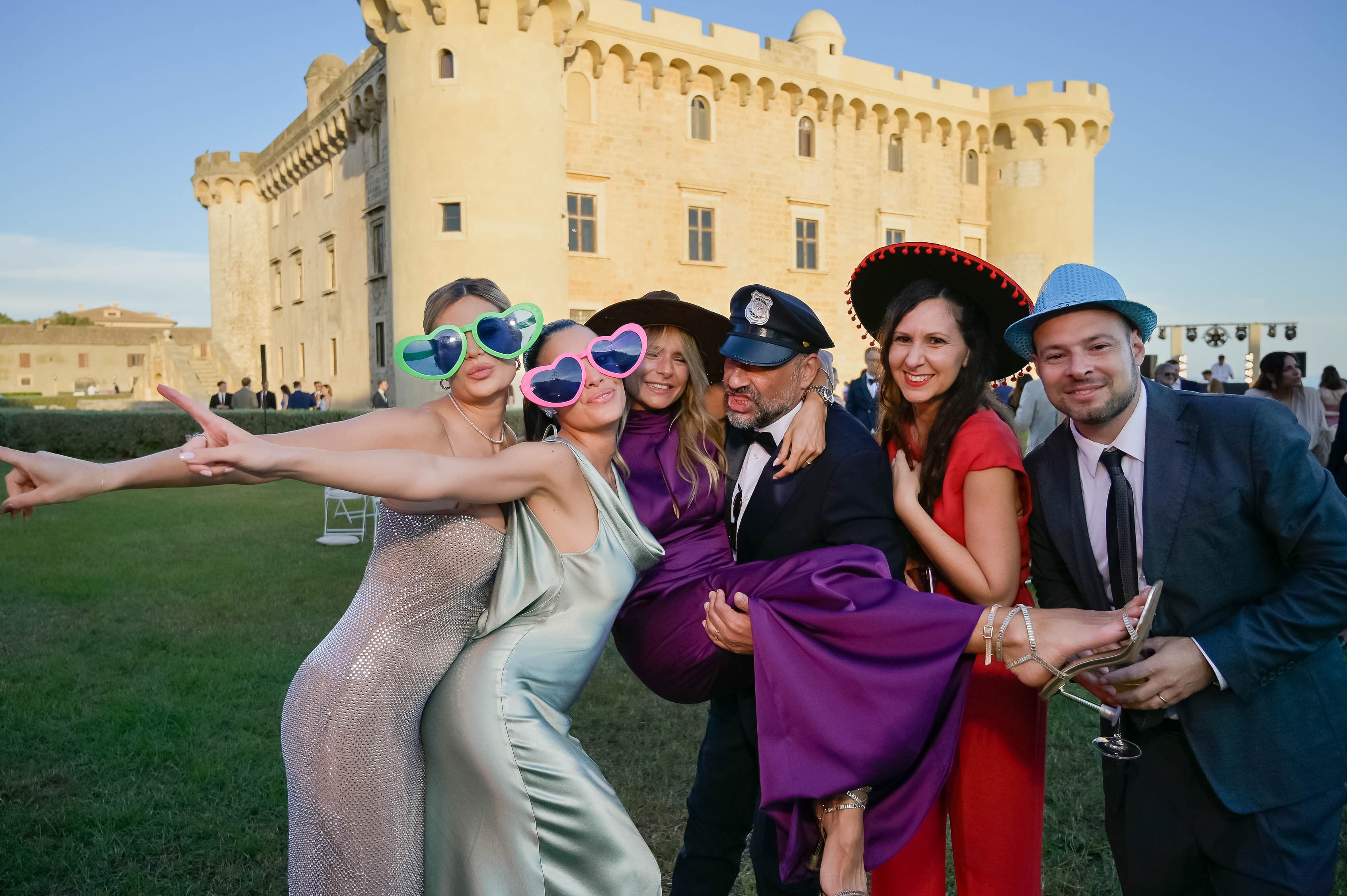 Gruppo di invitati a un matrimonio di lusso a Roma si diverte al photo booth Pro Selfie con cappelli e occhialoni davanti a un castello al tramonto